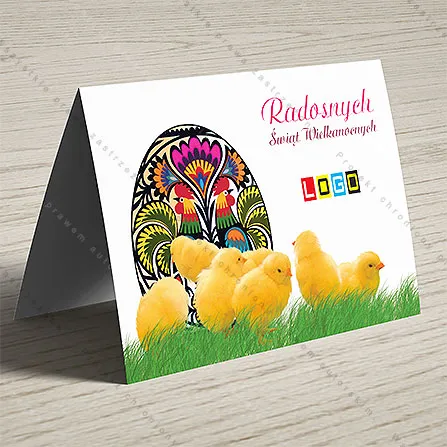Czarna pisanka w kolorowe kwiaty i dwoma kogutami, przed nią żółte pisklęta w trawie, z prawej malinowy napis